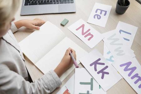 Un garçon écrit sur un cahier aidé par des fiches montrant des lettres minuscules et majuscules.